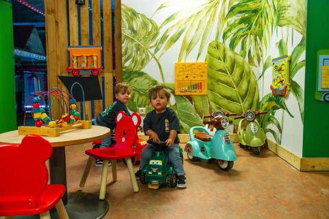 Twee jonge kinderen spelen in een kleurrijke speelkamer van een lodge met speelgoed en scooters.