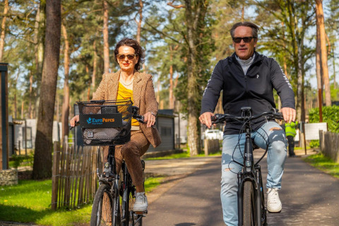 Twee volwassenen fietsen door een glamping accommodatie tussen de bomen, allebei met een zonnebril.