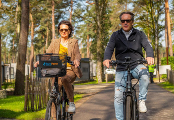 Twee volwassenen fietsen samen door een glamping verblijf tussen de bomen, beiden met zonnebril op.