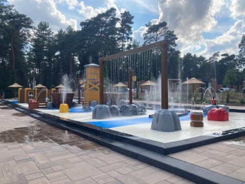 Aire de jeux aquatiques près d’un lodge, entourée d’arbres et de parasols, avec jets d’eau et jeux.