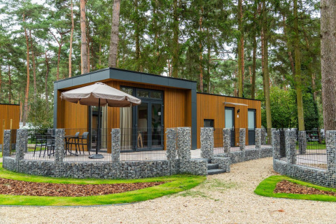 Cabina glamping nella foresta con pannelli in legno moderno, terrazza arredata e recinzione in pietra.