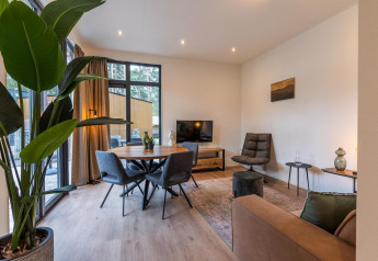 Moderne glamping-opholdsstue med store vinduer, spisebord, grøn plante og komfortable møbler.