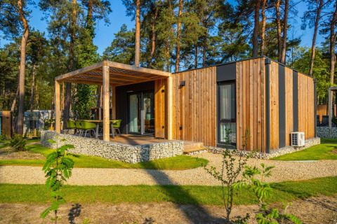 Cabaña moderna de madera con terraza cubierta y sillas verdes, ubicada en un entorno boscoso.