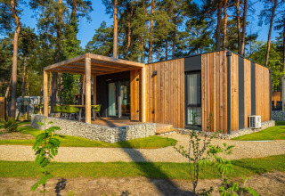 Cabaña moderna de madera con terraza cubierta y sillas verdes, ubicada en un entorno boscoso.
