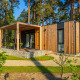 Moderne træhytte med terrasse og grønne stole i skovområde, ideel til afslappende udendørs ophold.