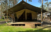 Glamping tent met houten terras en zwarte stoelen midden in een bosrijke omgeving op een zonnige dag.