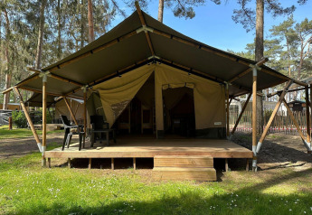 Tente de glamping avec terrasse en bois et chaises noires, située dans une forêt par une journée ensoleillée.