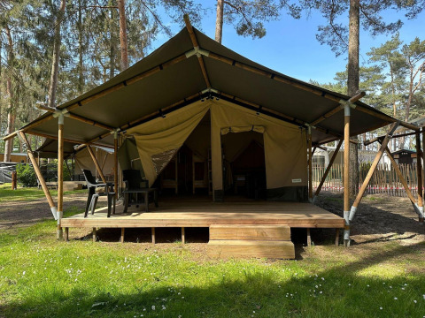 Tente de glamping avec terrasse en bois et chaises noires, située dans une forêt par une journée ensoleillée.