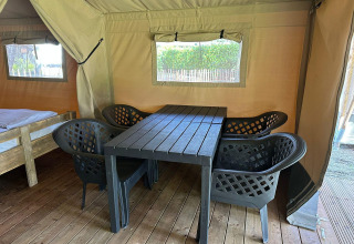Interior de tienda glamping con suelo de madera, cuatro sillas negras y mesa junto a una cama sencilla.