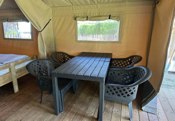 Interior de tienda glamping con suelo de madera, cuatro sillas negras y mesa junto a una cama sencilla.