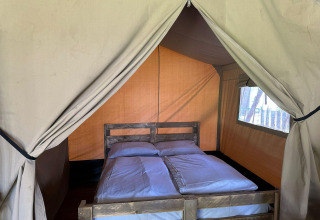 Interior de una tienda glamping con una cama doble, ropa de cama blanca y un marco de madera visto de frente.