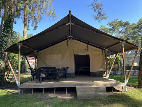 Grande tenda glamping con veranda in legno e sedie all'aperto, immersa tra alberi in un campeggio tranquillo.