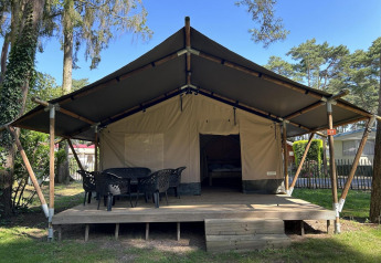 Groot glamping-tent met houten veranda en tuinstoelen, omgeven door bomen in een rustige campingomgeving.