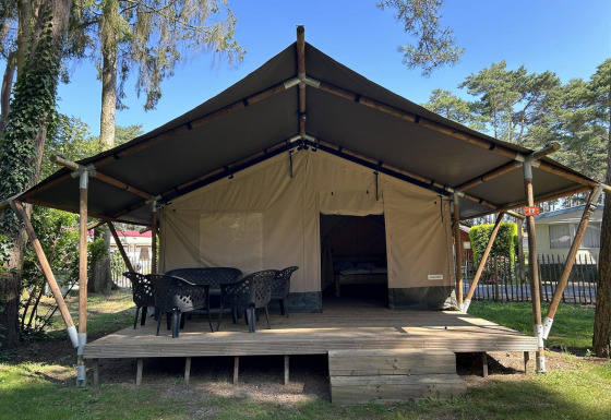 Grande tenda glamping con veranda in legno e sedie all'aperto, immersa tra alberi in un campeggio tranquillo.
