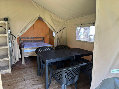 Interior de una tienda glamping con cama de madera, mesa y sillas plásticas, y suelo de madera iluminado.