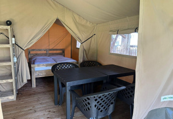 Interior de una tienda glamping con cama de madera, mesa y sillas plásticas, y suelo de madera iluminado.