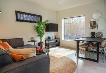Moderne lille stue i Forest Cottage ved Hoge Kempen, Belgien, med sofa, spisebord og store vinduer.