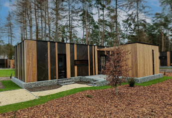 Moderne lille skovhytte i Hoge Kempen, Belgien, med træ- og stenfacade omgivet af nåletræer.