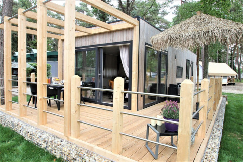 Kempen cottage lodge med moderne træterrasse, store vinduer og græsplæne omgivet af skov.