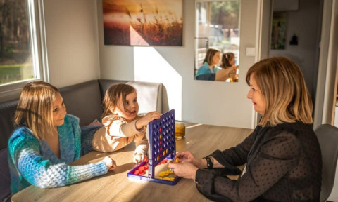 Tres personas juegan un juego de mesa en una mesa en Kempen cottage, Hoge Kempen, Bélgica, con luz solar.