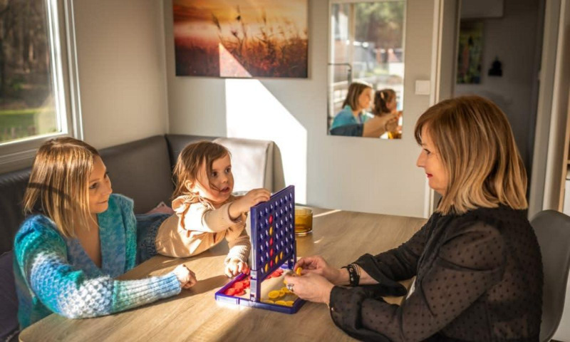 Tres personas juegan un juego de mesa en una mesa en Kempen cottage, Hoge Kempen, Bélgica, con luz solar.