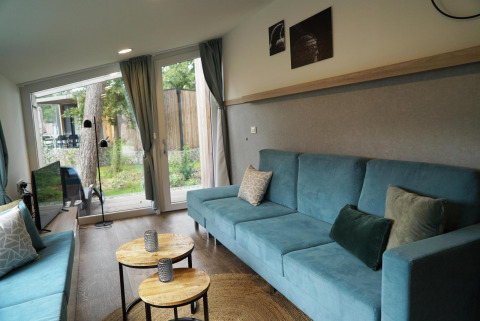 Salón moderno de una tiny house en Hoge Kempen, Bélgica, con sofás azules, mesas redondas y ventanas grandes.
