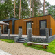 Moderne lodge i skoven med træbeklædning, terrasse, parasol og stenmur, omgivet af grøn natur.