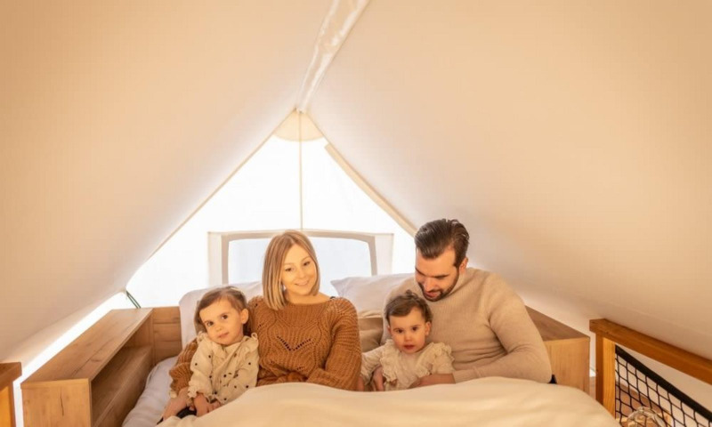 Eine Familie sitzt zusammen im Bett in der Papendaal Lodge im Hoge Kempen, Belgien, bei weichem Licht.