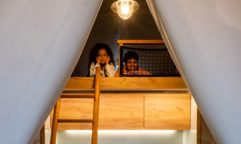 Dos niños sonríen en un altillo de Papendaal Lodge en Hoge Kempen, Bélgica, iluminados por una lámpara cálida.