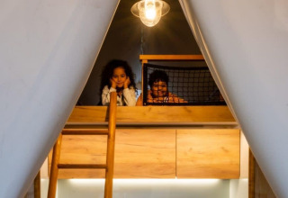 Dos niños sonríen en un altillo de Papendaal Lodge en Hoge Kempen, Bélgica, iluminados por una lámpara cálida.