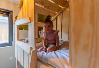 Niña pequeña sentada en la cama en una cabaña infantil en Hoge Kempen, Bélgica, con decoración de madera.