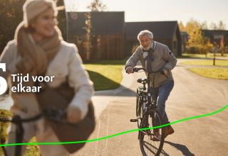 Koppel op leeftijd fietst op zonnig pad bij moderne huisjes in Unbrick One | Sauna and Pool lodge, fijne tijd samen.