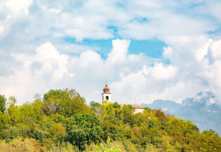 Kerktoren tussen bomen bij Camping Al Lago di Lago in Veneto, Italië, onder een bewolkte blauwe hemel.