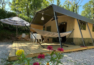 Safari tent Base Lodge with wooden deck, hammock, parasol, flowers in foreground, and green trees behind.