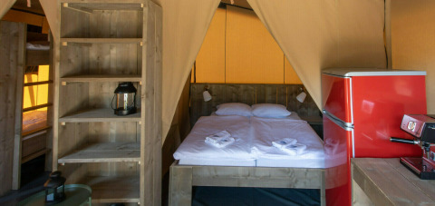Interior de la tienda safari Base Lodge en Camping Al Lago di Lago, Italia, con cama, estantería y nevera roja.