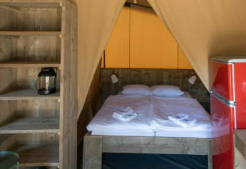 Interior de la tienda safari Base Lodge en Camping Al Lago di Lago, Italia, con cama, estantería y nevera roja.