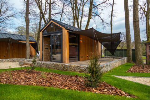 Modern tiny house with wood exterior, large windows, and shaded terrace surrounded by trees and grass.
