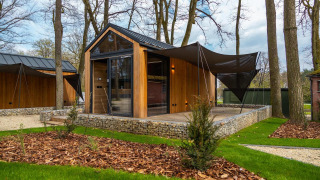 Moderne tiny house med træbeklædning, store vinduer og udendørs terrasse omgivet af træer og grønt.