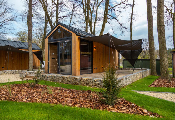 Modern tiny house with wood exterior, large windows, and shaded terrace surrounded by trees and grass.