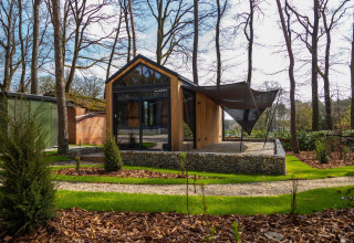 Casa pequeña moderna en el bosque de Hoge Kempen, Bélgica, con fachada de madera y terraza sombreada.
