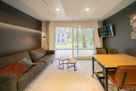 Sala luminosa en la cabaña Thor, Hoge Kempen, Bélgica, con sofá, TV, mesa y vista al bosque exterior.