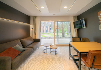 Sala luminosa en la cabaña Thor, Hoge Kempen, Bélgica, con sofá, TV, mesa y vista al bosque exterior.