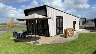 Just Nature Light lodge met hedendaags ontwerp, terras met stoelen, parasol en houtstapel op het grasveld.