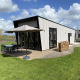 Just Nature Light-lodge med moderne design, udendørs siddeplads, parasol og stablet brænde på græsplæne.