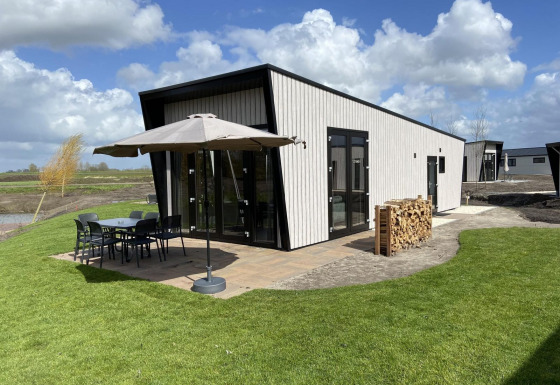 Just Nature Light lodge met hedendaags ontwerp, terras met stoelen, parasol en houtstapel op het grasveld.