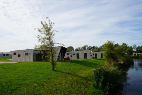 Lodge Just Nature moderno junto a un lago, rodeado de césped y árboles bajo un cielo luminoso.