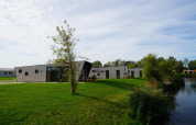 Moderne Just Nature lodge ved en sø, omgivet af grønne plæner og træer under en blå himmel.