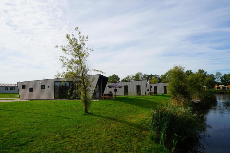 Lodge moderne Just Nature au bord d’un lac, entouré de pelouse verte et d’arbres sous un ciel clair.