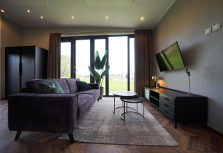 Sala moderna en la cabaña Just Nature de las Islas IJssel, Países Bajos, con sofá, TV y ventanales al jardín.