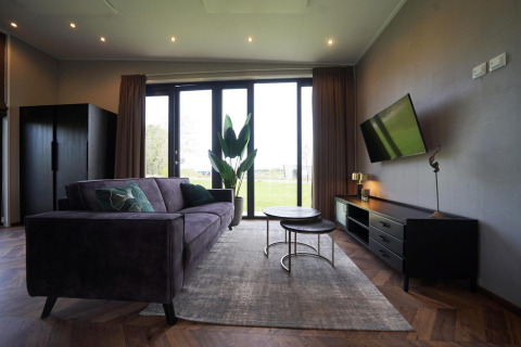 Sala moderna en la cabaña Just Nature de las Islas IJssel, Países Bajos, con sofá, TV y ventanales al jardín.